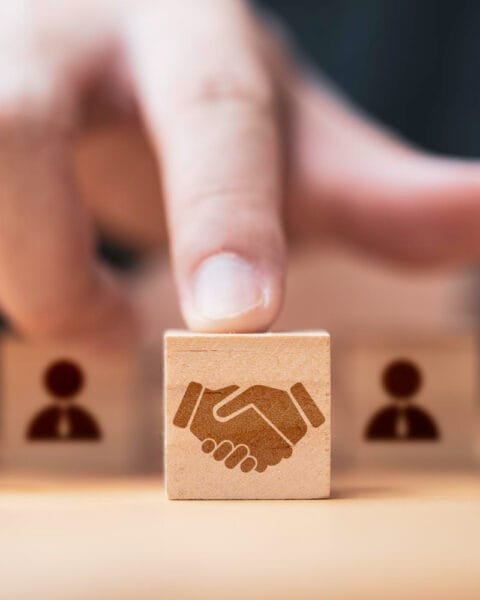 A finger points at a wooden block featuring a handshake icon to represent Asia-Pacific M&A Market Analysis, surrounded by blocks with person silhouettes, symbolizing partnership.