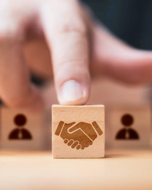 3396 A finger points at a wooden block featuring a handshake icon to represent Asia-Pacific M&A Market Analysis, surrounded by blocks with person silhouettes, symbolizing partnership.