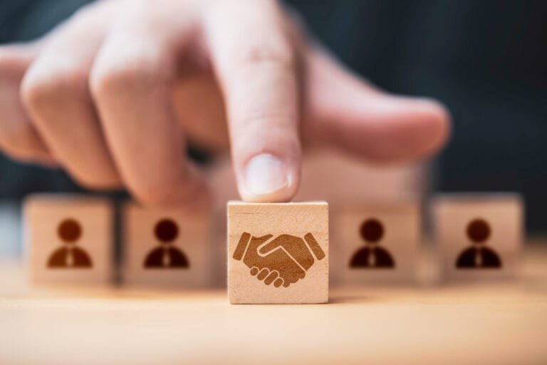 A finger points at a wooden block featuring a handshake icon to represent Asia-Pacific M&A Market Analysis, surrounded by blocks with person silhouettes, symbolizing partnership.