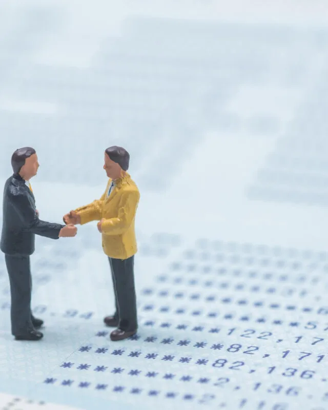 Asia M&A Trends 2025 - Two miniature figures in business attire shake hands over a background of financial documents, symbolizing a business agreement.