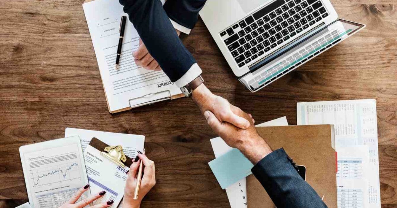Asia M&A $4T Market Forecast: Two business professionals shake hands over a wooden desk filled with documents and a laptop, signifying a deal or agreement.