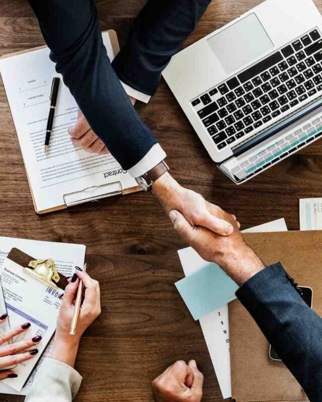 Asia M&A $4T Market Forecast: Two business professionals shake hands over a wooden desk filled with documents and a laptop, signifying a deal or agreement.