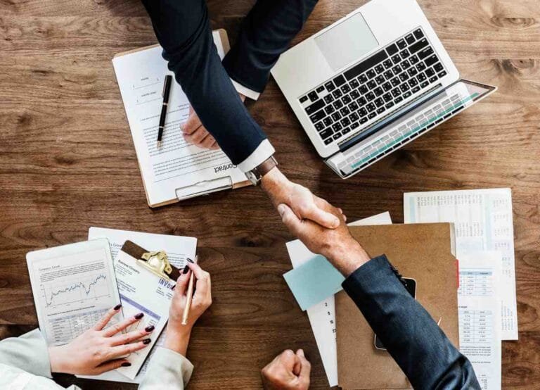 Asia M&A $4T Market Forecast: Two business professionals shake hands over a wooden desk filled with documents and a laptop, signifying a deal or agreement.
