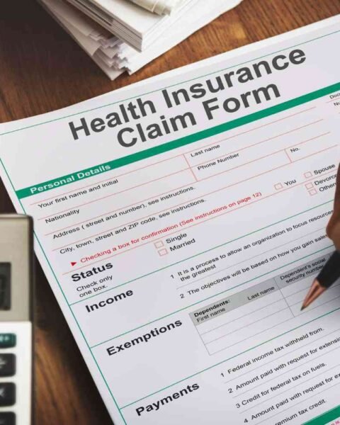 M&A Insurance Uptake Rises: A hand holding a pen fills out a health insurance claim form beside a calculator and stacks of documents on a wooden table.