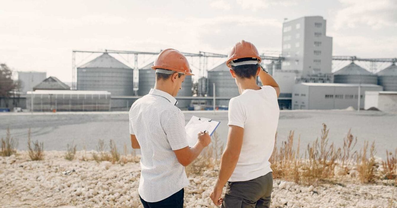 Asia Infrastructure Fundraising: Two construction workers in hard hats observe a large industrial facility, one holding a clipboard in a gravelly terrain.