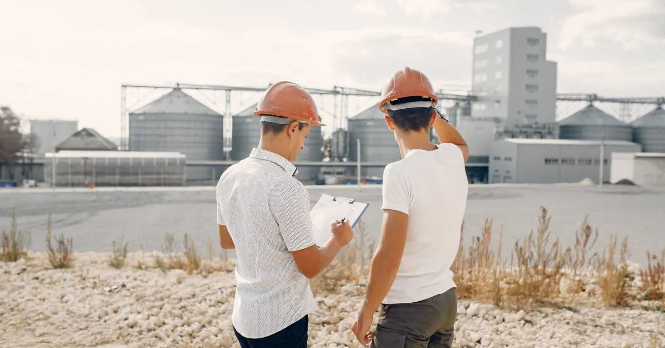 35333 Asia Infrastructure Fundraising: Two construction workers in hard hats observe a large industrial facility, one holding a clipboard in a gravelly terrain.
