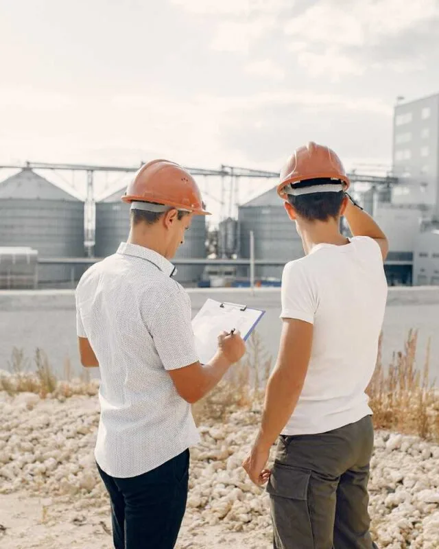 35333 Asia Infrastructure Fundraising: Two construction workers in hard hats observe a large industrial facility, one holding a clipboard in a gravelly terrain.