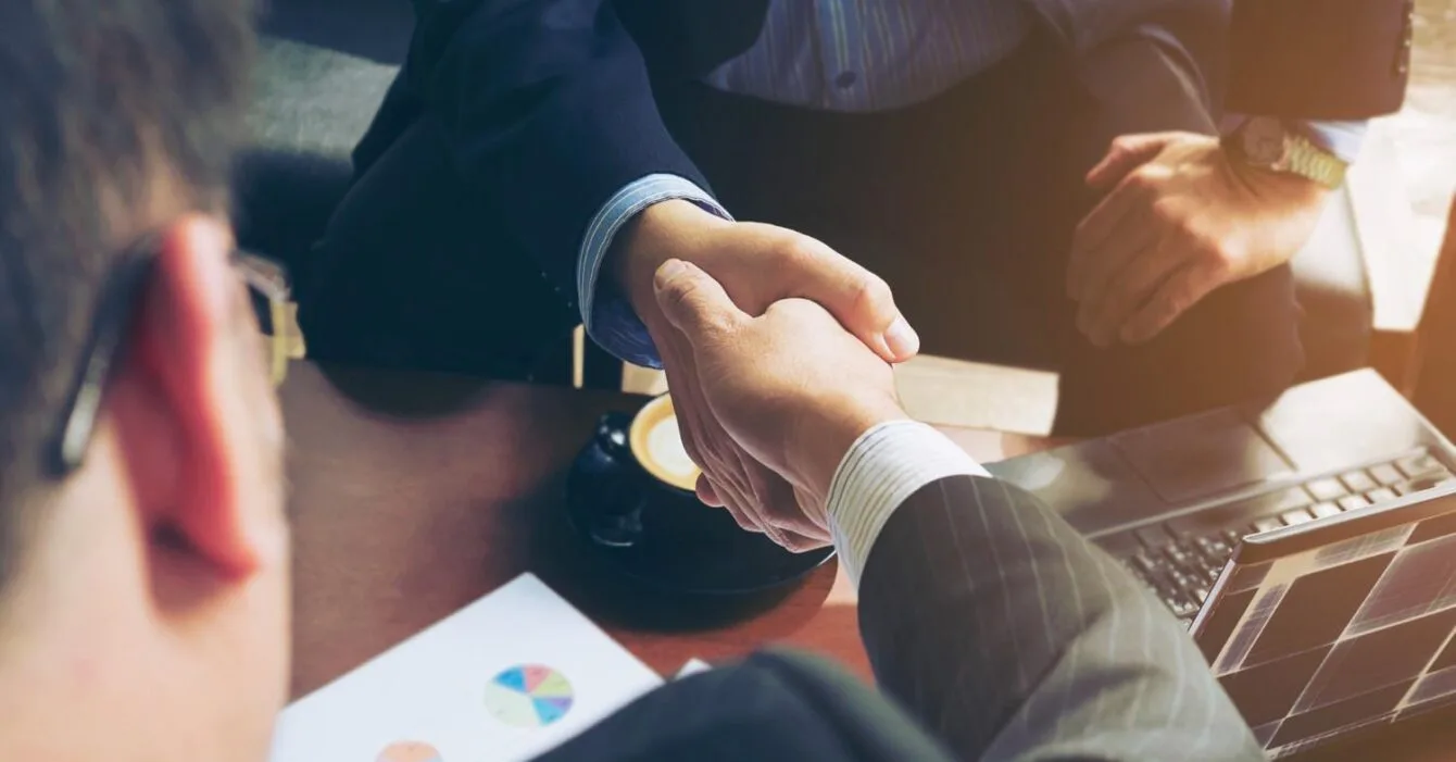 6402 Asia-Pacific M&A Value Growth: Two businessmen shake hands across a desk with a laptop and coffee, signifying a successful agreement or partnership.