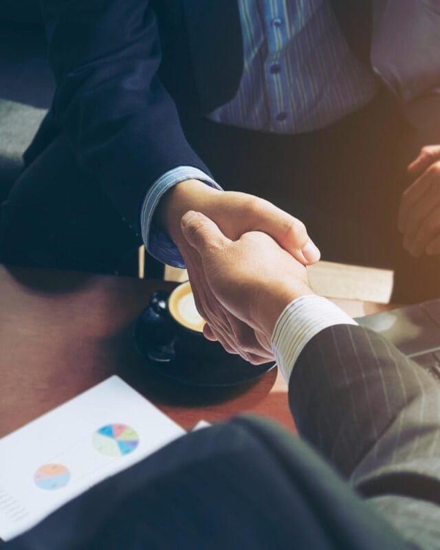 Asia-Pacific M&A Value Growth: Two businessmen shake hands across a desk with a laptop and coffee, signifying a successful agreement or partnership.