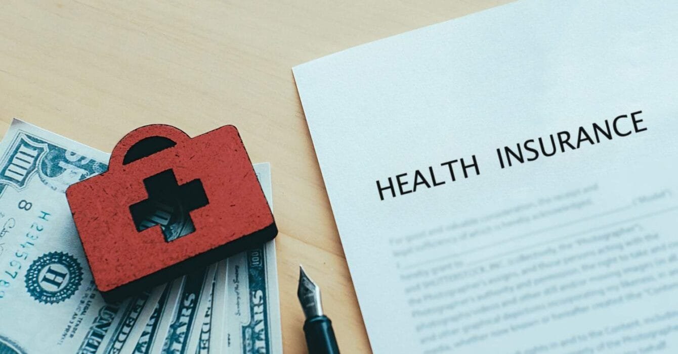 Asia M&A Insurance Trends: A red medical bag sits atop dollar bills next to a health insurance document and a fountain pen on a wooden surface.