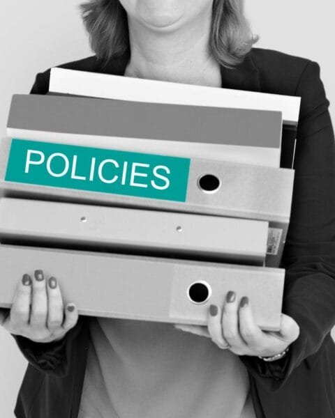 Asia M&A Regulatory Delays: A woman in a blazer holds several binders labeled "POLICIES," symbolizing organization and management of policy documents.