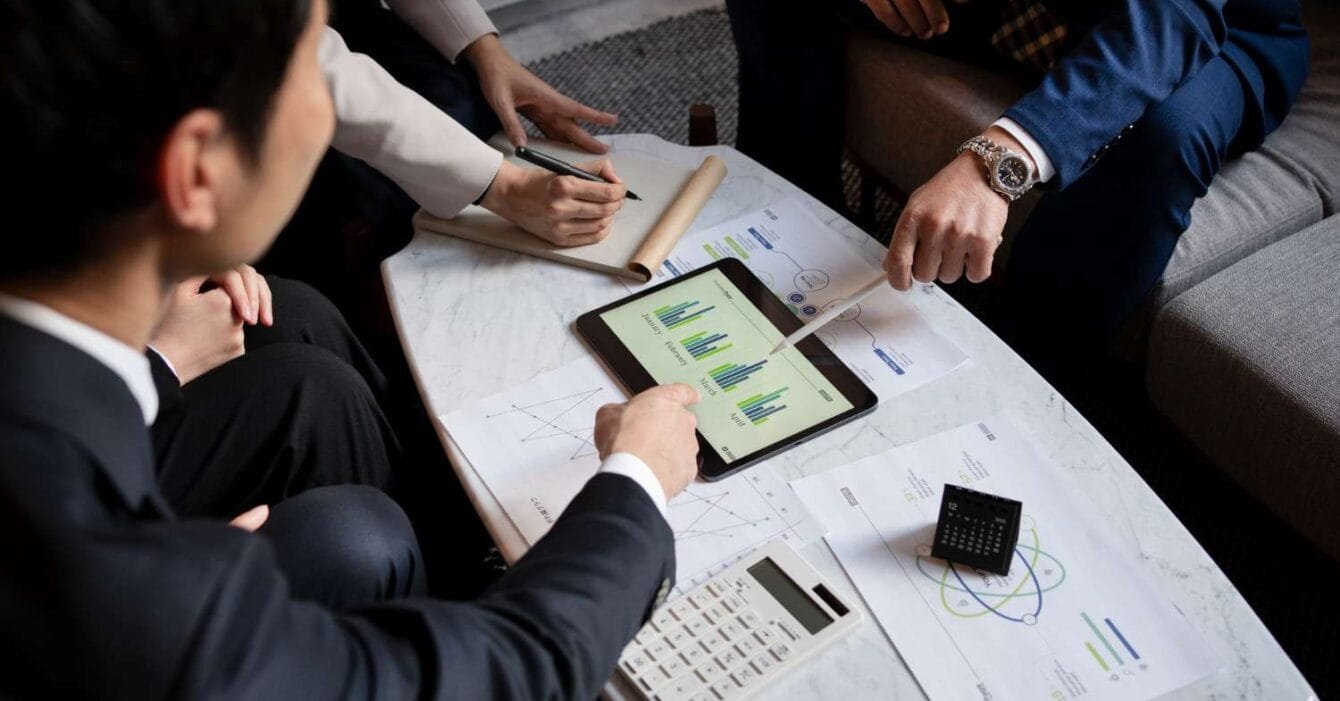 2150103555 Asia M&A Private Equity Momentum: A group of people discussing charts on a tablet, with documents, a calculator, and pens on a marble table in a modern setting.
