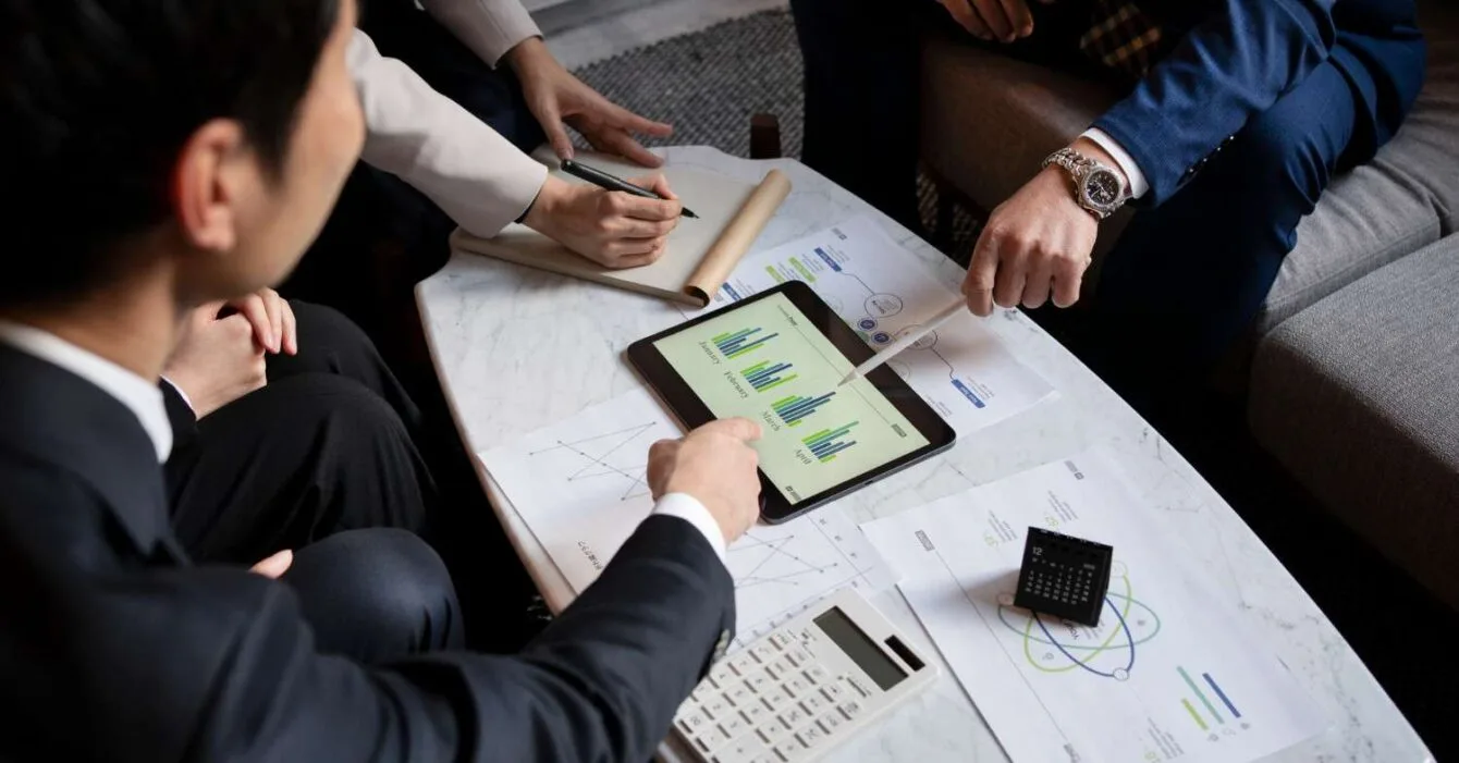 Asia M&A Private Equity Momentum: A group of people discussing charts on a tablet, with documents, a calculator, and pens on a marble table in a modern setting.
