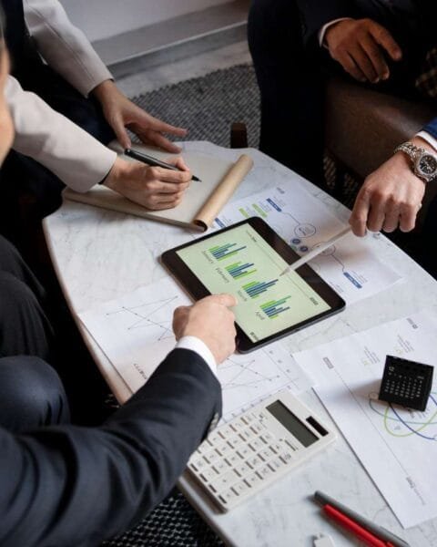Asia M&A Private Equity Momentum: A group of people discussing charts on a tablet, with documents, a calculator, and pens on a marble table in a modern setting.