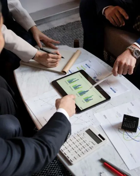 Asia M&A Private Equity Momentum: A group of people discussing charts on a tablet, with documents, a calculator, and pens on a marble table in a modern setting.