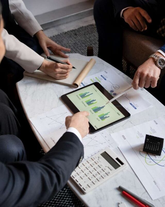 2150103555 Asia M&A Private Equity Momentum: A group of people discussing charts on a tablet, with documents, a calculator, and pens on a marble table in a modern setting.