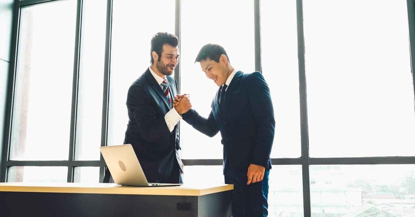 getty-images-2tor9SbDs_Q-unsplash Asia M&A Deal Value Growth: Two business professionals shake hands in front of a large window, with a laptop on the desk beside them, symbolizing a successful agreement.