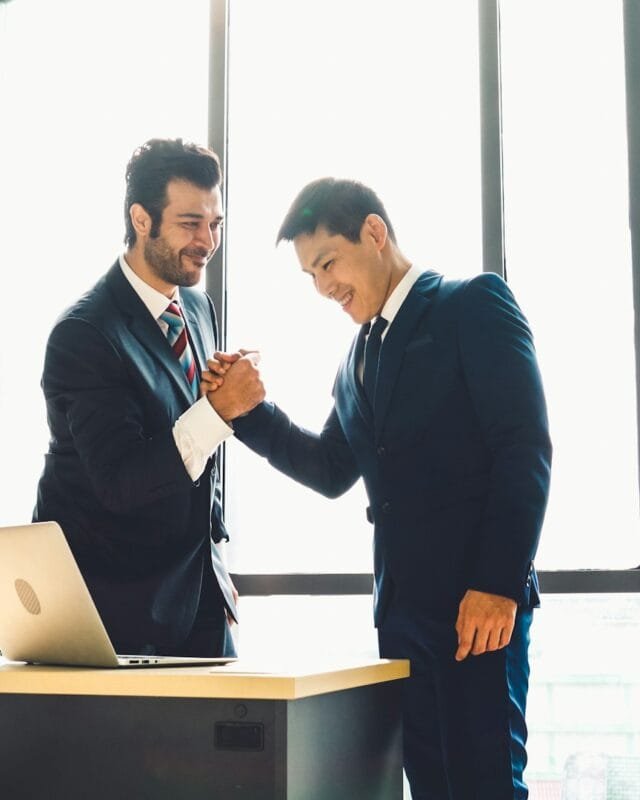 Asia M&A Deal Value Growth: Two business professionals shake hands in front of a large window, with a laptop on the desk beside them, symbolizing a successful agreement.