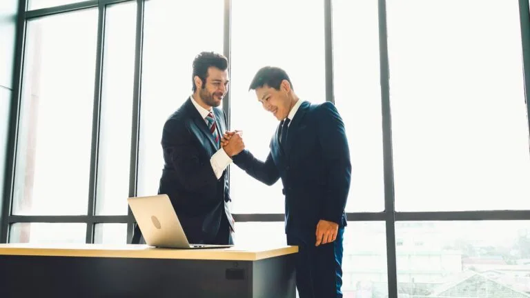 Asia M&A Deal Value Growth: Two business professionals shake hands in front of a large window, with a laptop on the desk beside them, symbolizing a successful agreement.