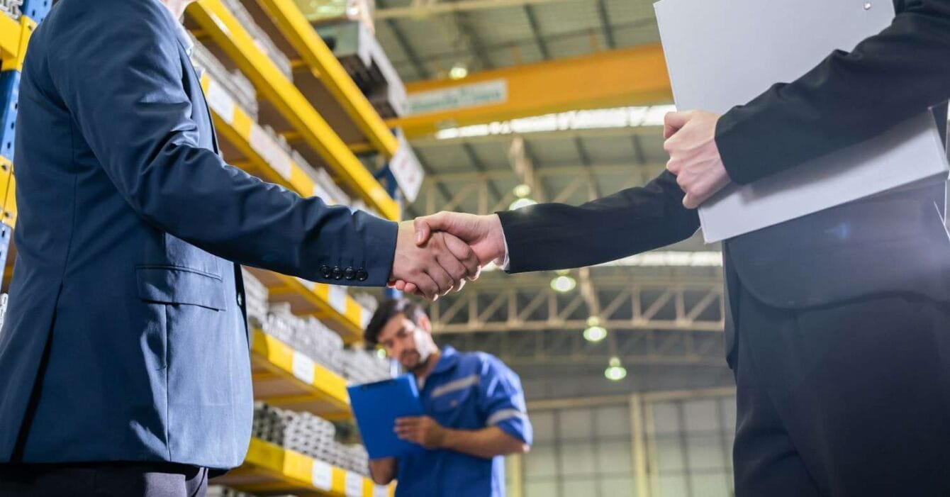 getty-images-F307ZBFSIoA-unsplash Asia M&A Optimism 2025: Two professionals shake hands in a warehouse setting, with shelves stocked in the background and a third worker holding a clipboard nearby.