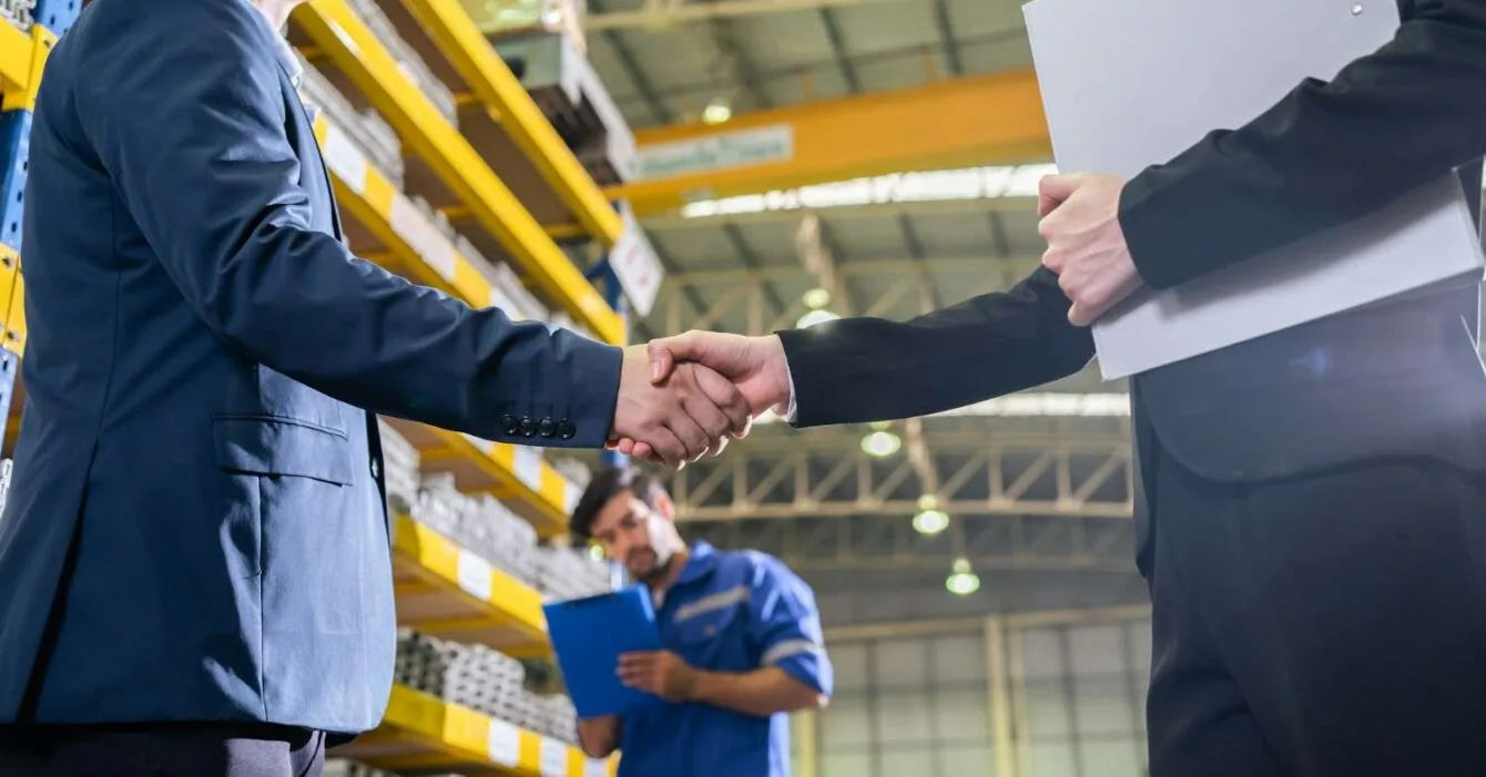 Asia M&A Optimism 2025: Two professionals shake hands in a warehouse setting, with shelves stocked in the background and a third worker holding a clipboard nearby.