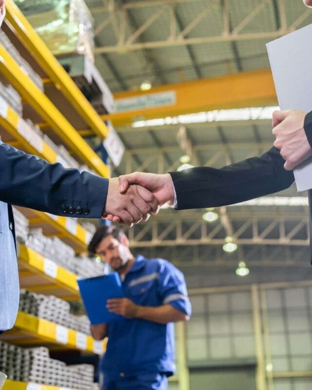 Asia M&A Optimism 2025: Two professionals shake hands in a warehouse setting, with shelves stocked in the background and a third worker holding a clipboard nearby.