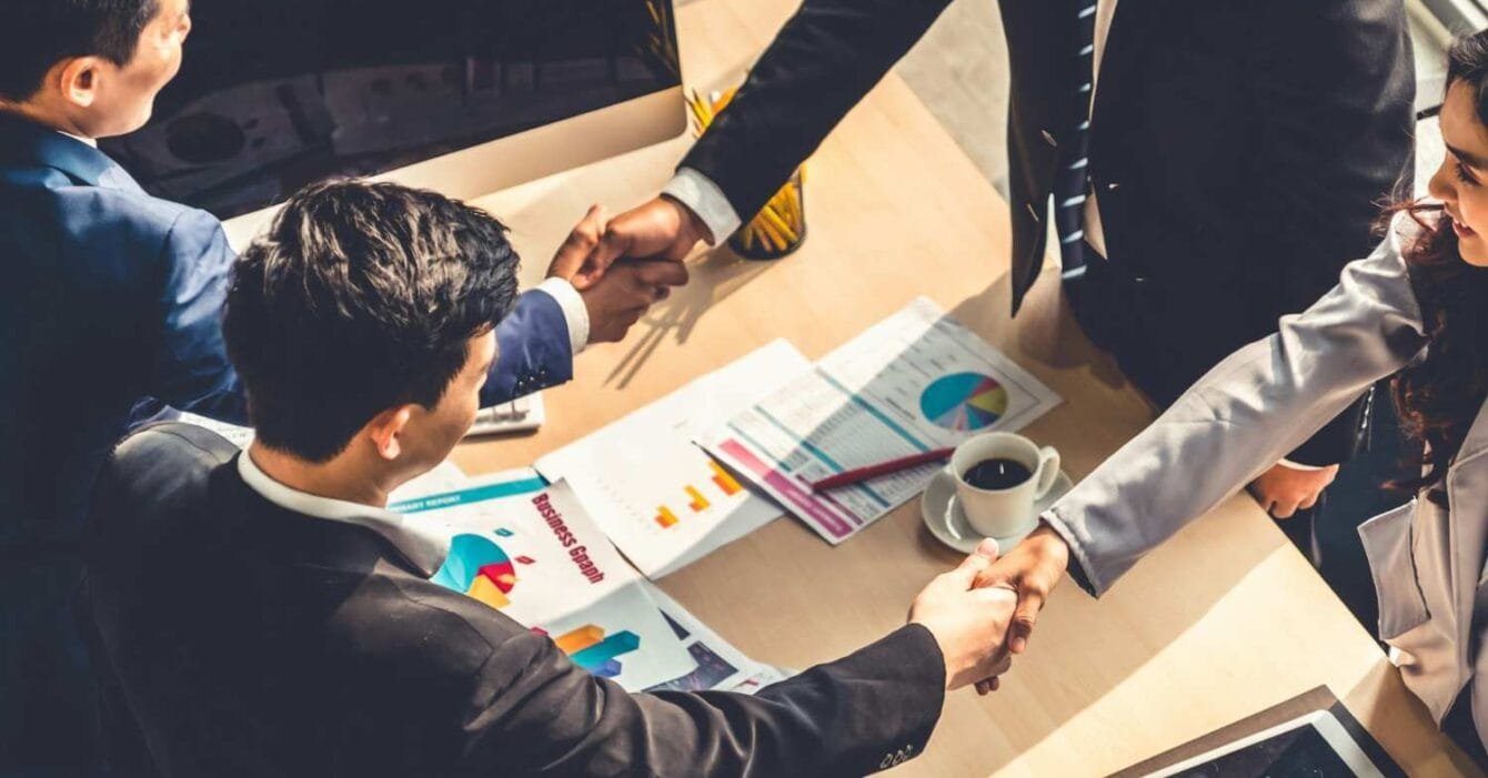 A group of professionals in business attire shake hands over a table with documents and a coffee cup, symbolizing a partnership.