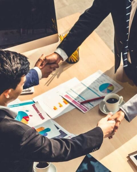 A group of professionals in business attire shake hands over a table with documents and a coffee cup, symbolizing a partnership.