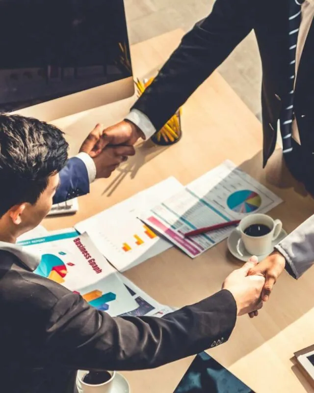 getty-images-JQaCvV46IXU-unsplash A group of professionals in business attire shake hands over a table with documents and a coffee cup, symbolizing a partnership.