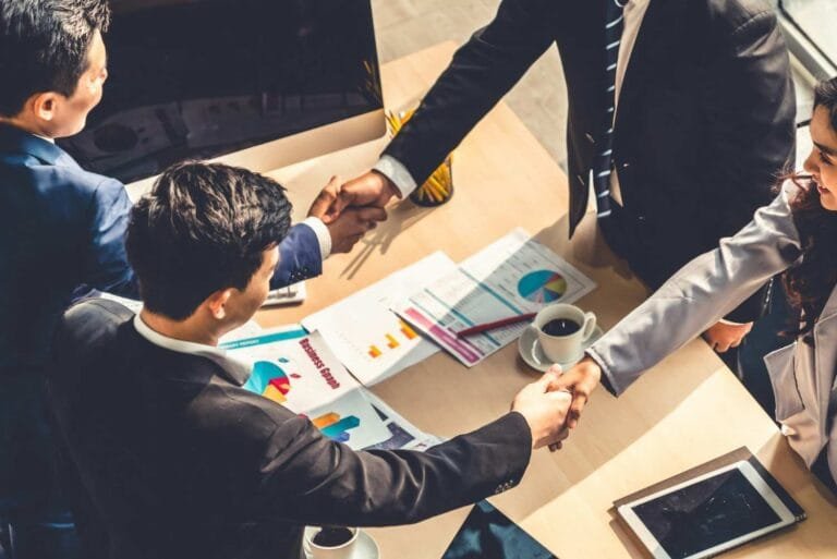 A group of professionals in business attire shake hands over a table with documents and a coffee cup, symbolizing a partnership.